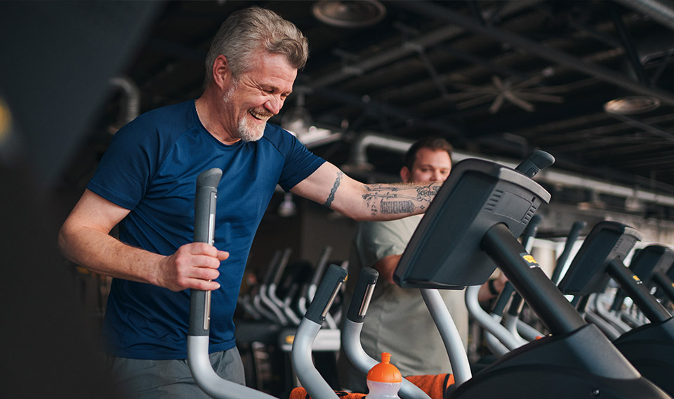 Imagen: Un hombre mayor entrena sonriendo en una elíptica en la zona de cardio. Al fondo se ve a otra persona entrenando.